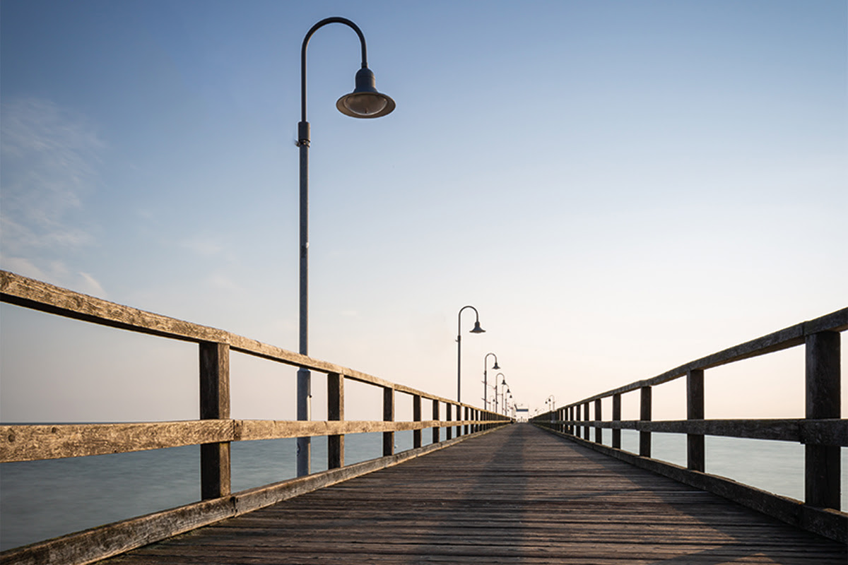 Holzseebrücke mit Laternen führt ins offene Meer bei klarem Himmel und ruhiger See.