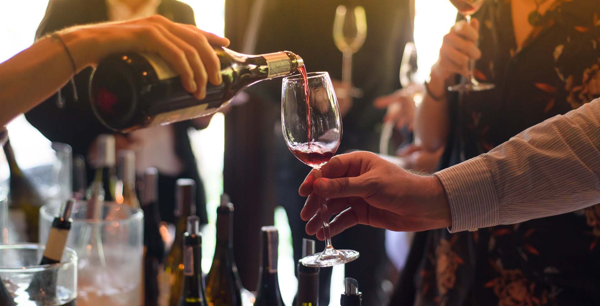 Person schenkt Rotwein aus einer Flasche in ein Glas, weitere Flaschen und Personen im Hintergrund.