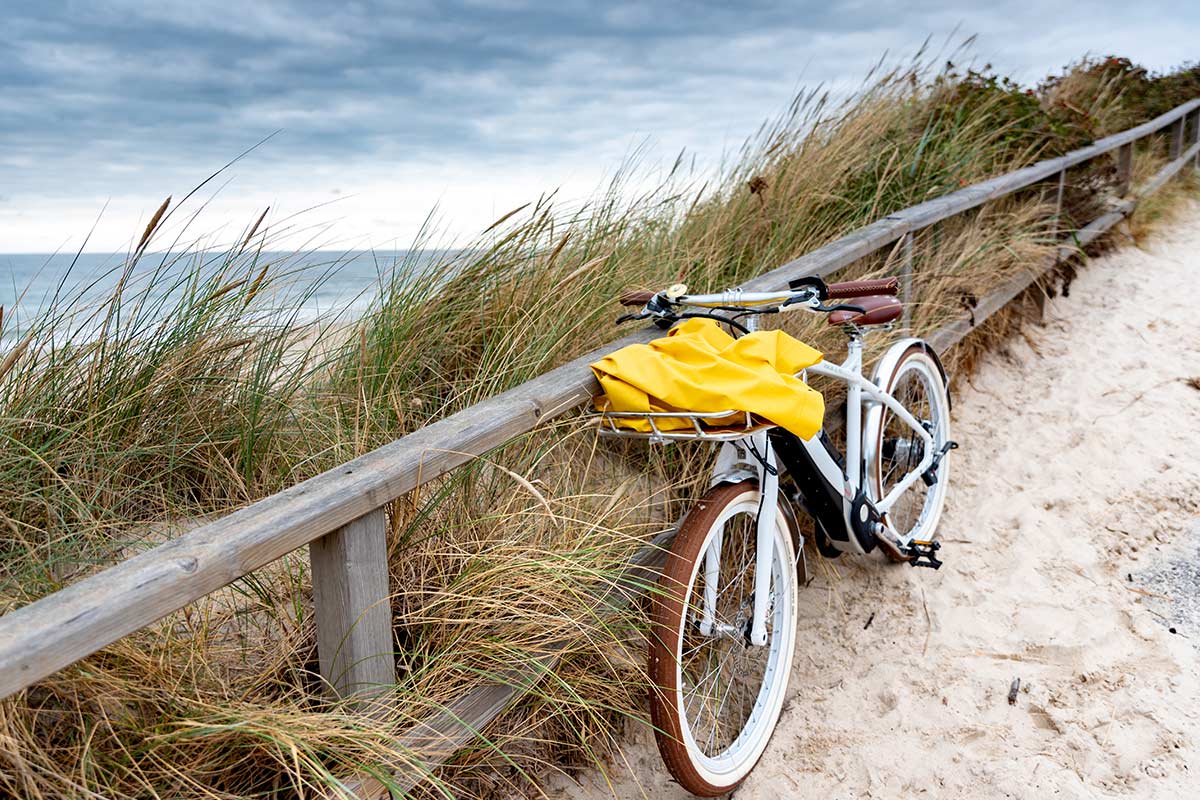 A bicycle with a yellow towel lies against a dune fence, the sandy path leads to the sea.