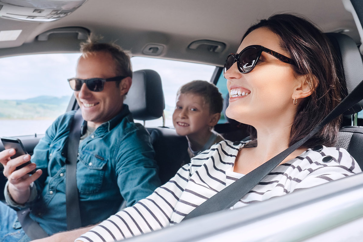 Family smiling while driving, father behind the wheel, mother and child in the back seat.