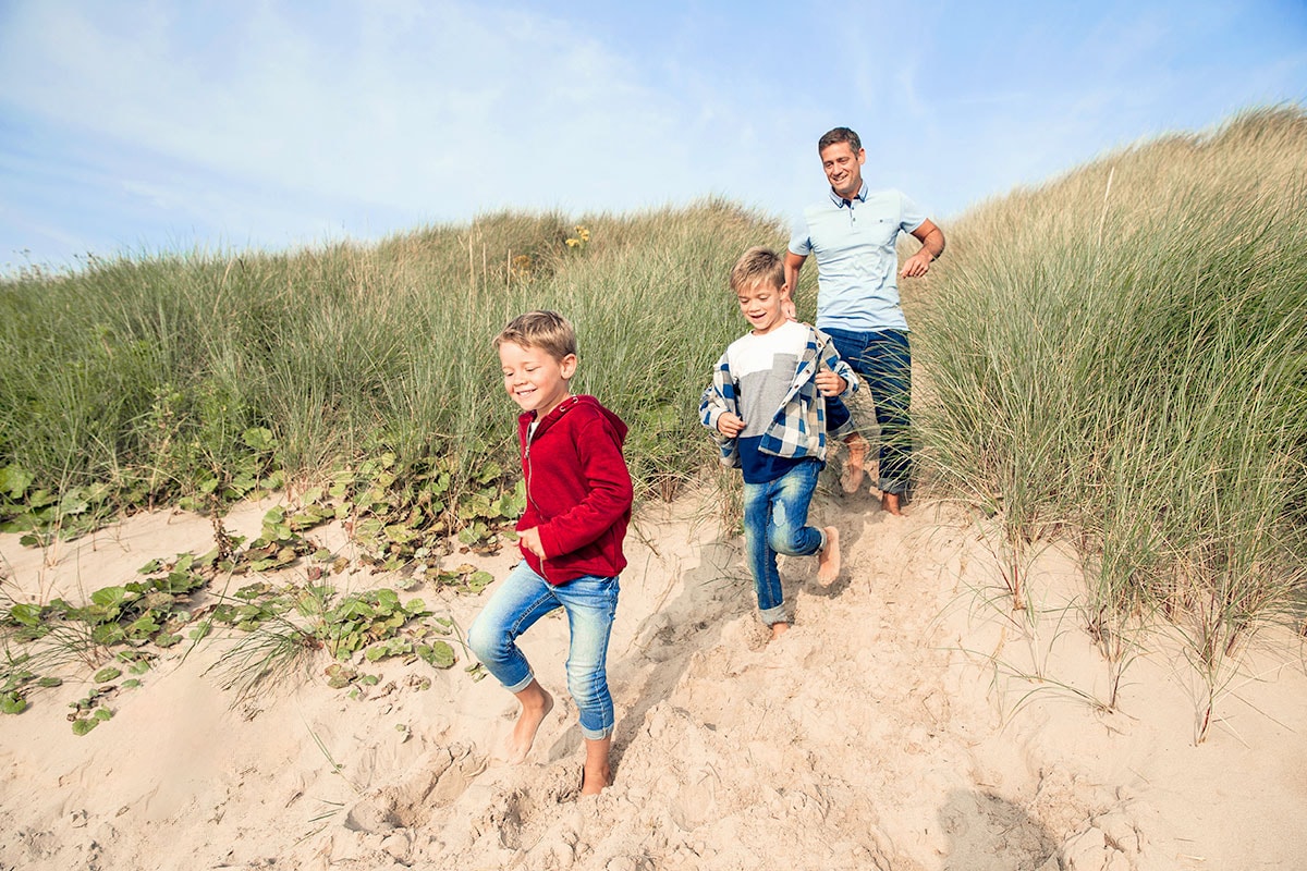 Father and two children walk barefoot through the dunes on the beach, everyone is smiling and enjoying the sunny day.
