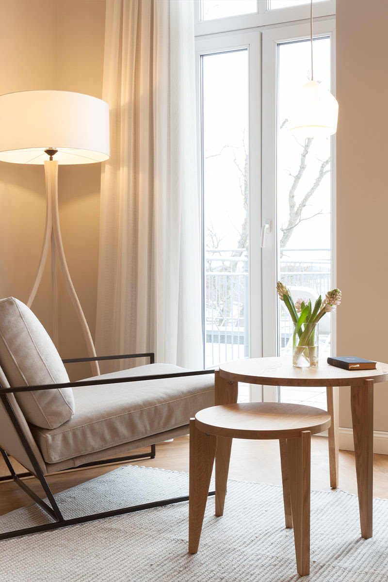 Armchair with cushions next to a small wooden table, floor lamp and flowers, window in the background.
