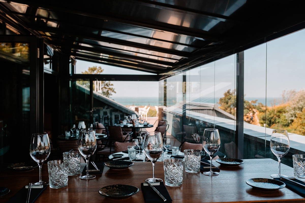 Set tables with glasses and plates in a modern glass-fronted restaurant with a sea view.