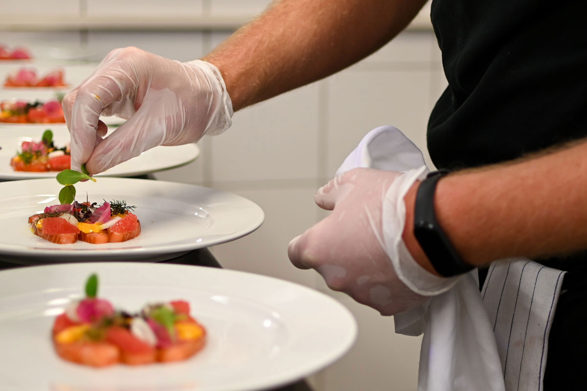 Chef wearing gloves artfully arranges a dish on a plate with more plates in a professional kitchen.