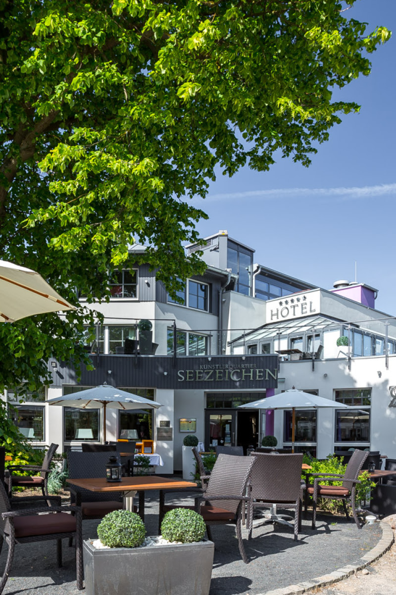 Exterior view of the Hotel Künstlerquartier Seezeichen with a cozy terrace under a large tree in sunny weather.