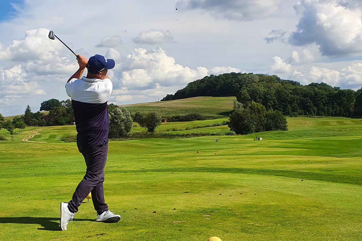 Golfer beim Abschlag auf einem Golfplatz, umgeben von grüner Landschaft und leicht bewölktem Himmel.
