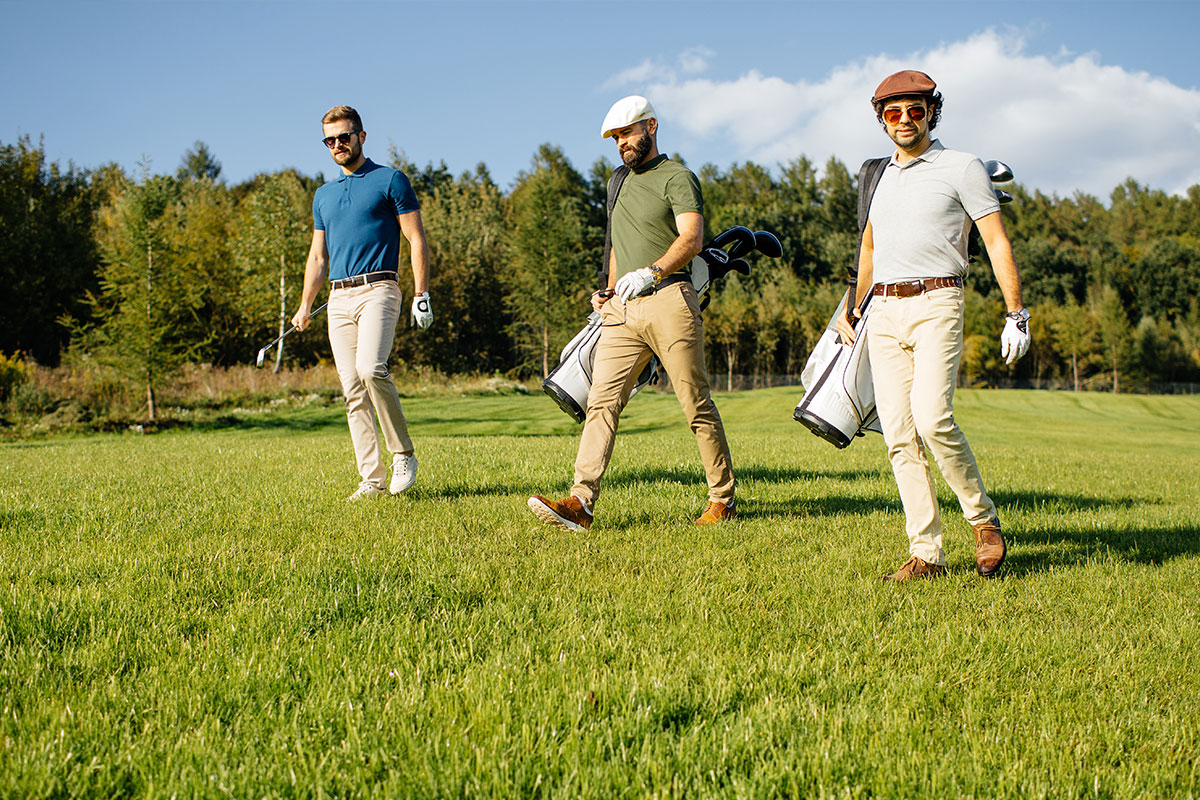 Drei Golfer gehen mit Golfschlägern und Taschen über das Fairway, umgeben von Natur und strahlendem Sonnenschein.