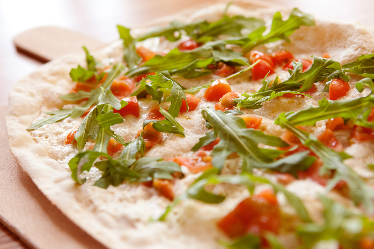 Nahaufnahme einer dünnen, knusprigen Pizza, belegt mit frischem Rucola und geschnittenen Tomaten.