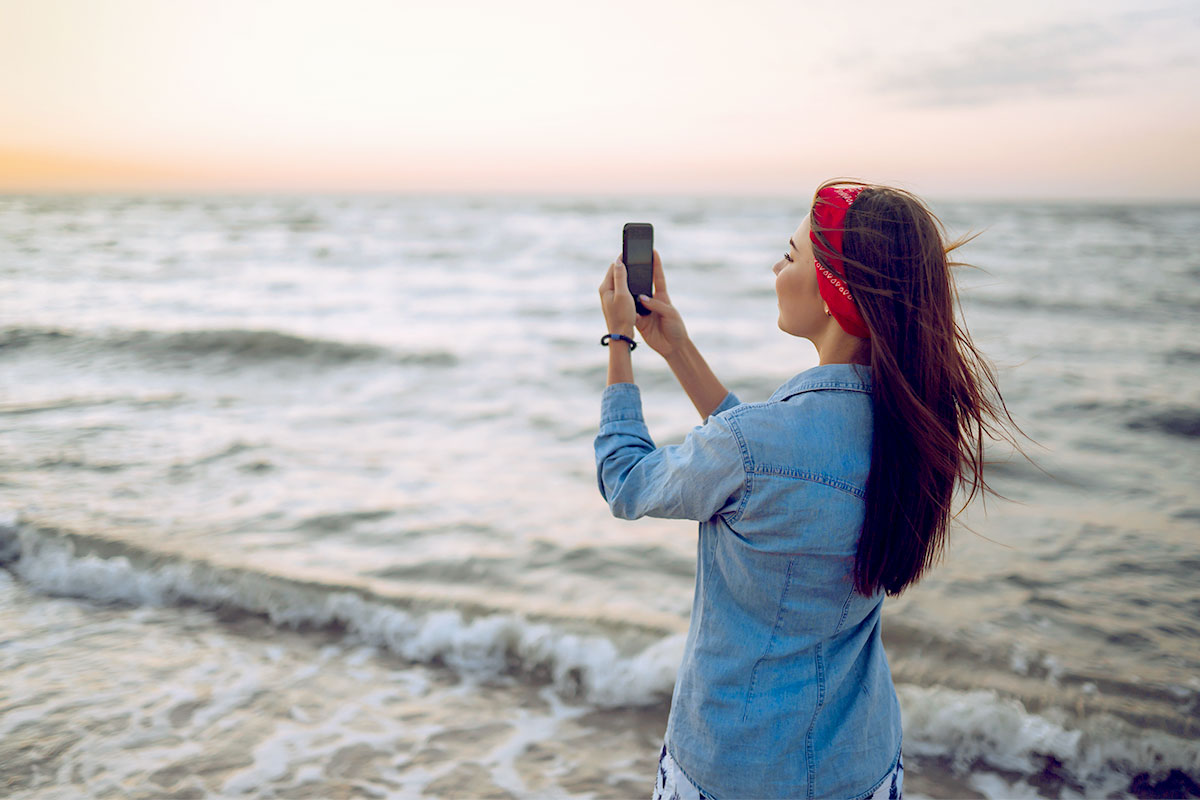 Frau am Strand, die mit ihrem Smartphone den Sonnenuntergang über dem Meer fotografiert, während der Wind ihr Haar weht.