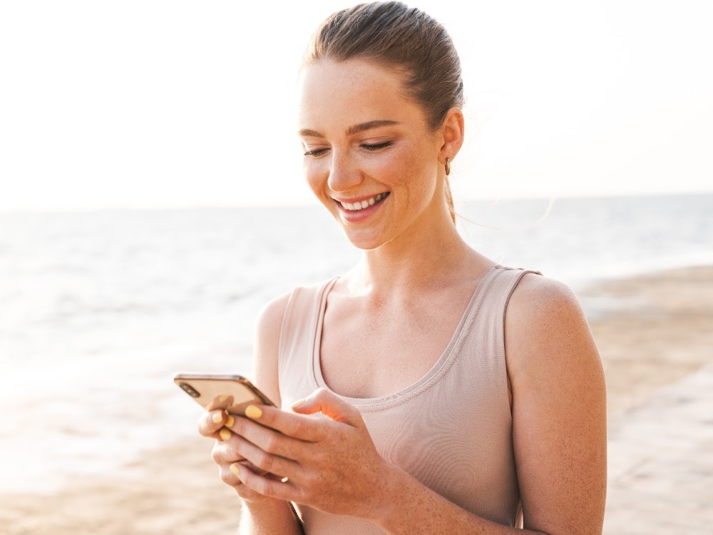 Eine Frau im ärmellosen Oberteil, die am Strand steht und auf ihr Smartphone lächelt.