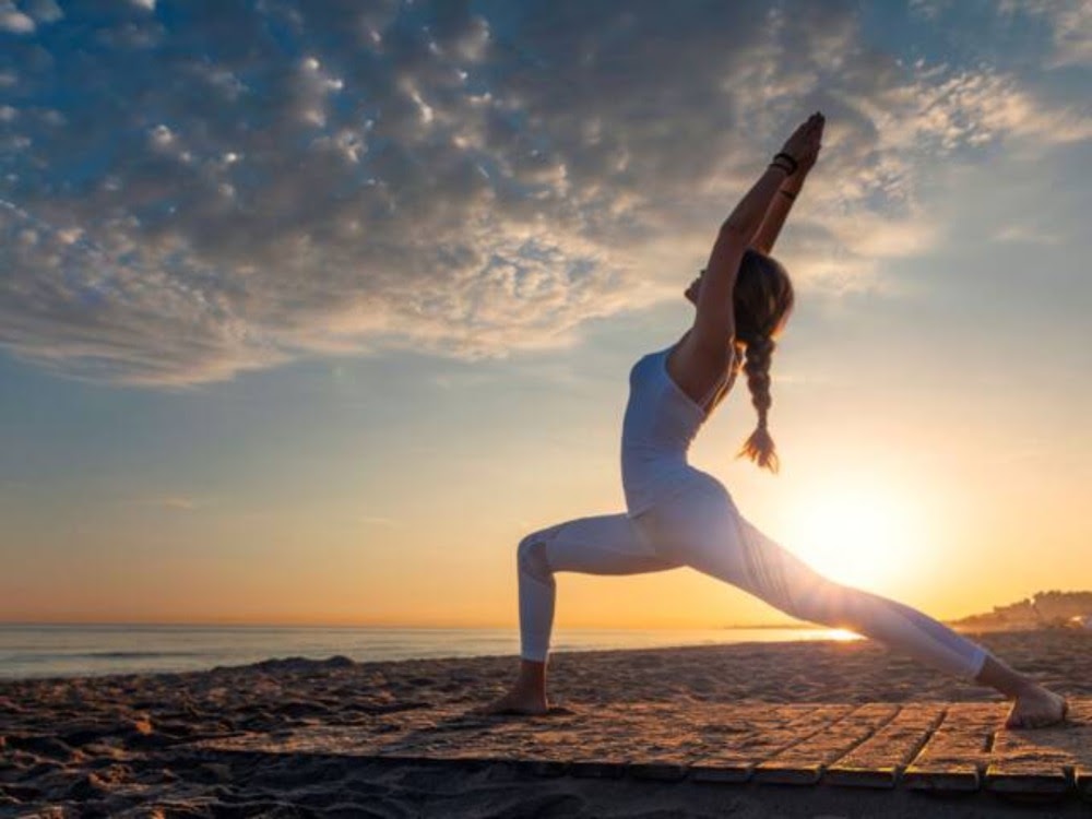 Frau in Yogapose am Strand bei Sonnenaufgang, sie streckt ihre Arme nach oben, der Himmel ist bewölkt.