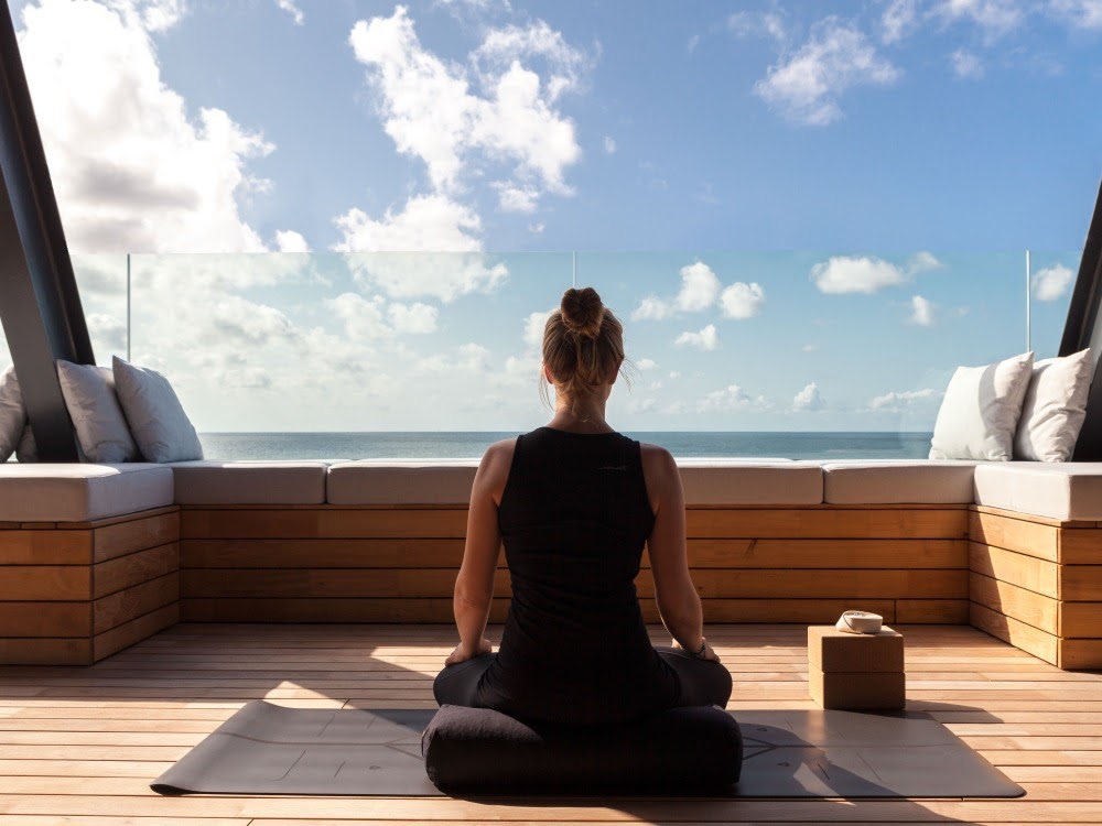 Person meditiert auf einer Holzterrasse vor einer Glaswand, die einen ungestörten Blick auf das Meer bietet.