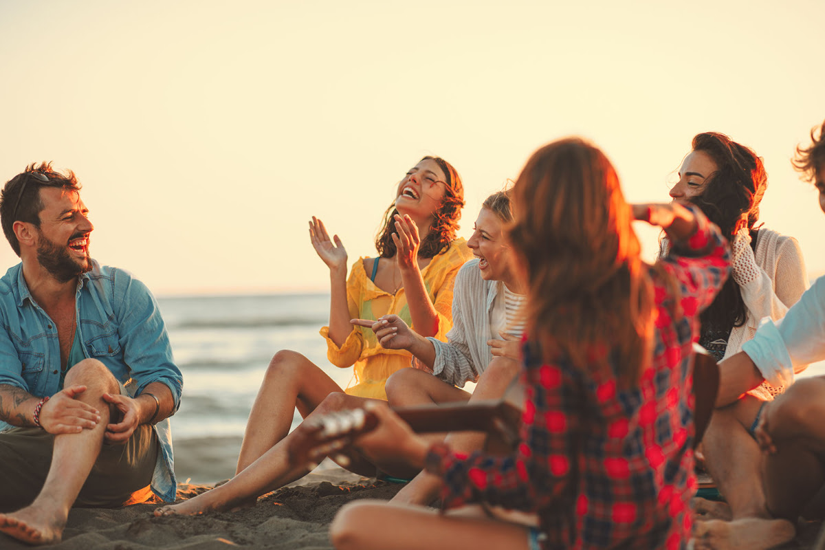 Gruppe junger Leute, die am Strand sitzt, lacht und Musik macht, während die Sonne untergeht.