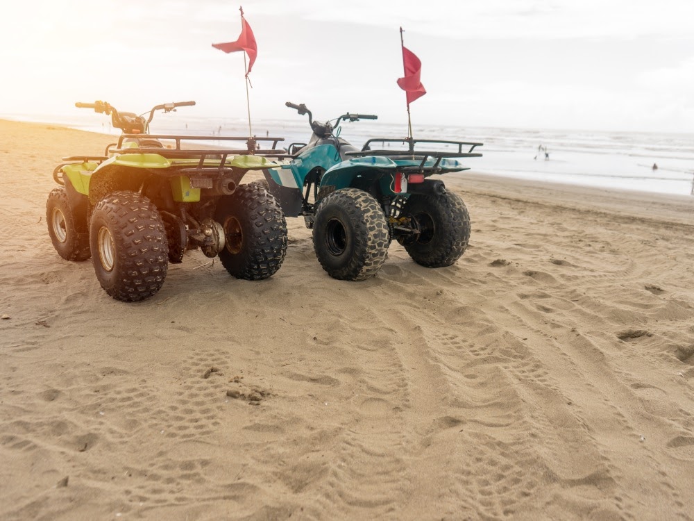 Zwei Quads mit roten Flaggen parken auf einem sonnigen Sandstrand mit tiefen Reifenabdrücken und Meerblick.