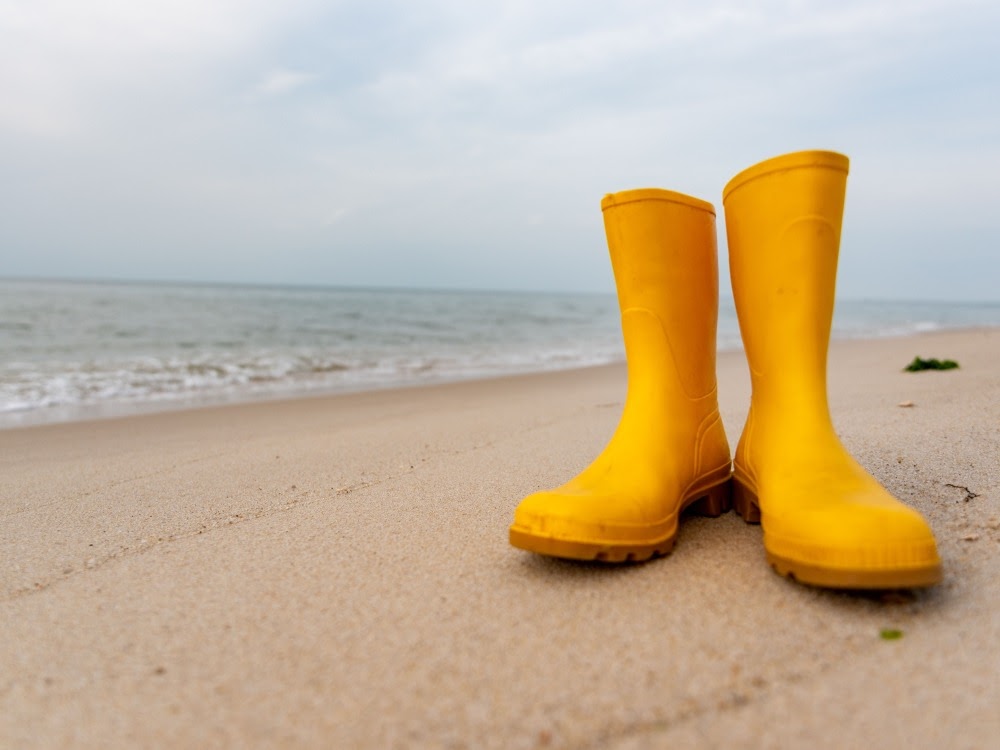 Gelbe Gummistiefel stehen auf einem sandigen Strand mit dem Meer im Hintergrund unter bewölktem Himmel.