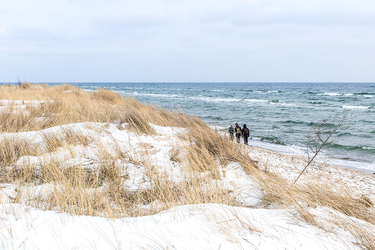 Menschen gehen an einem schneebedeckten Dünenstrand entlang das Meer mit kleinen Wellen im Hintergrund.