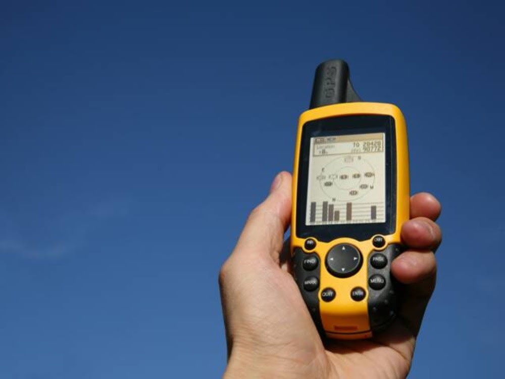 Eine Hand hält ein gelbes GPS-Gerät vor einem blauen Himmel mit Satellitensignalen auf dem Bildschirm.