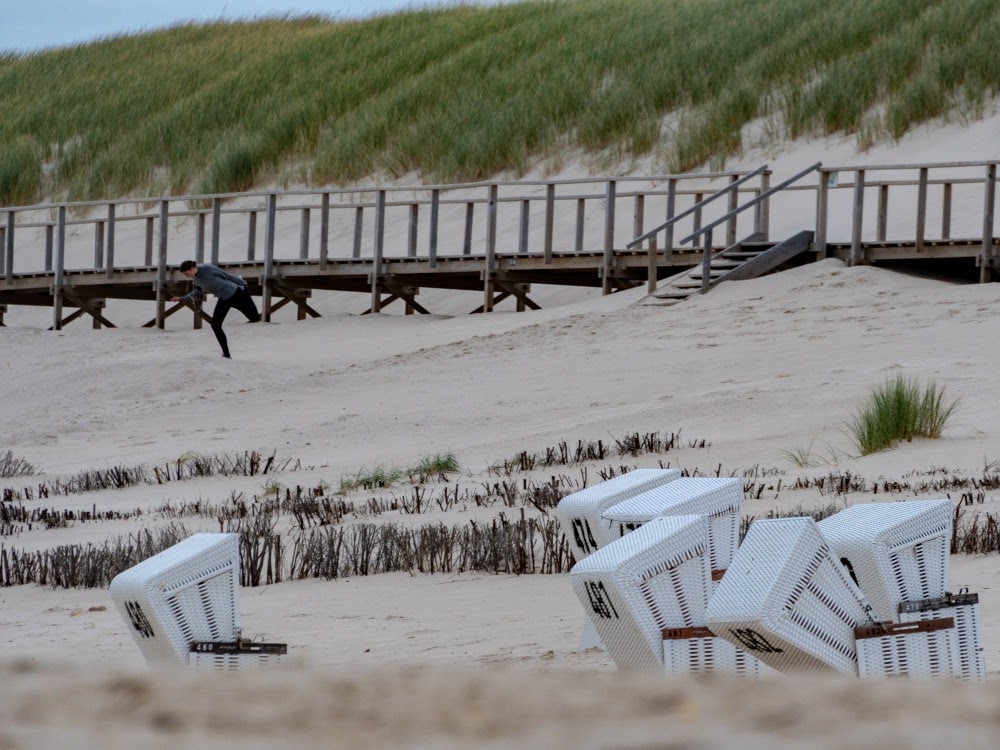 Weiße Strandkörbe an einem leeren Sandstrand, mit Holzsteg und grasbewachsenen Dünen im Hintergrund.