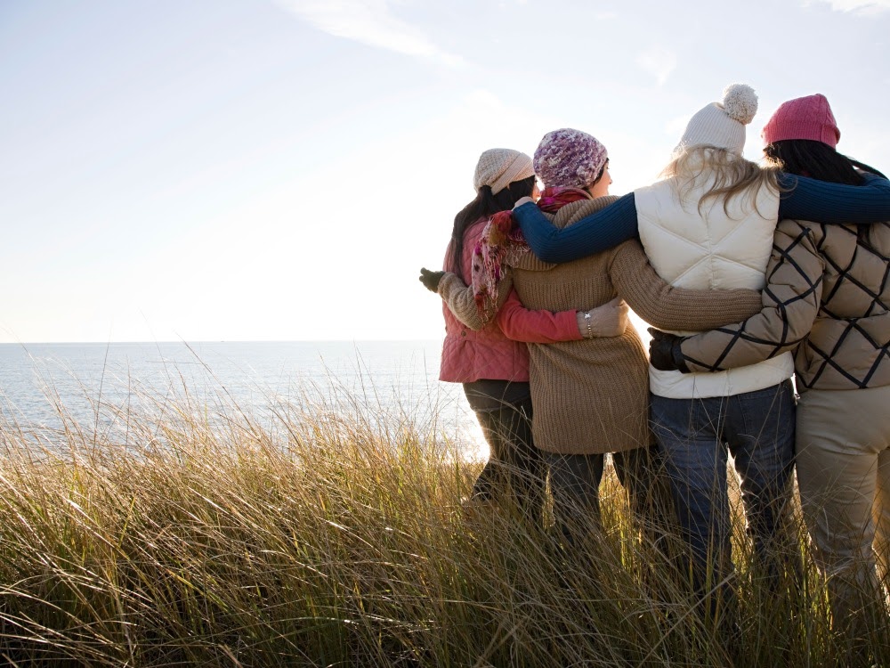 Vier Frauen stehen gemeinsam am Strand und blicken in die Ferne während sie sich umarmen.