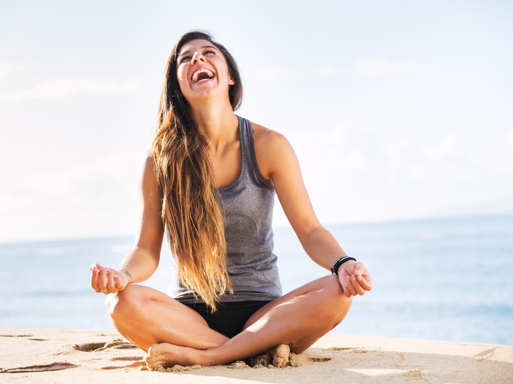 Frau sitzt lächelnd im Schneidersitz am Strand und macht Yoga während sie die Meeresbrise genießt.