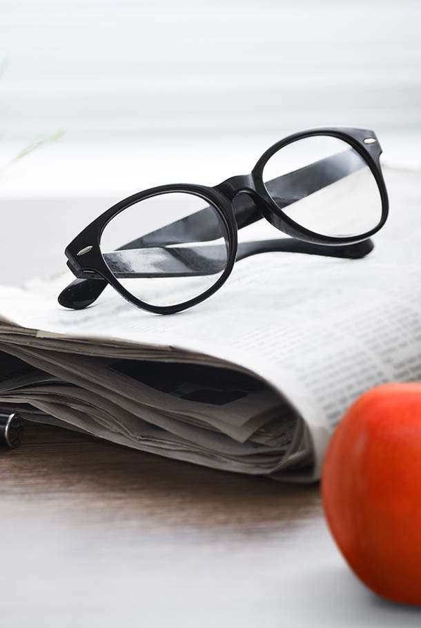 Black glasses on folded newspaper with an apple in the foreground, on a desk.