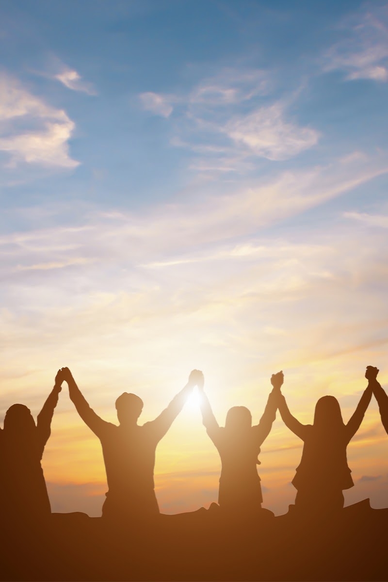 Personen stehen Hand in Hand und heben ihre Arme vor einem farbenprächtigen Sonnenuntergang.