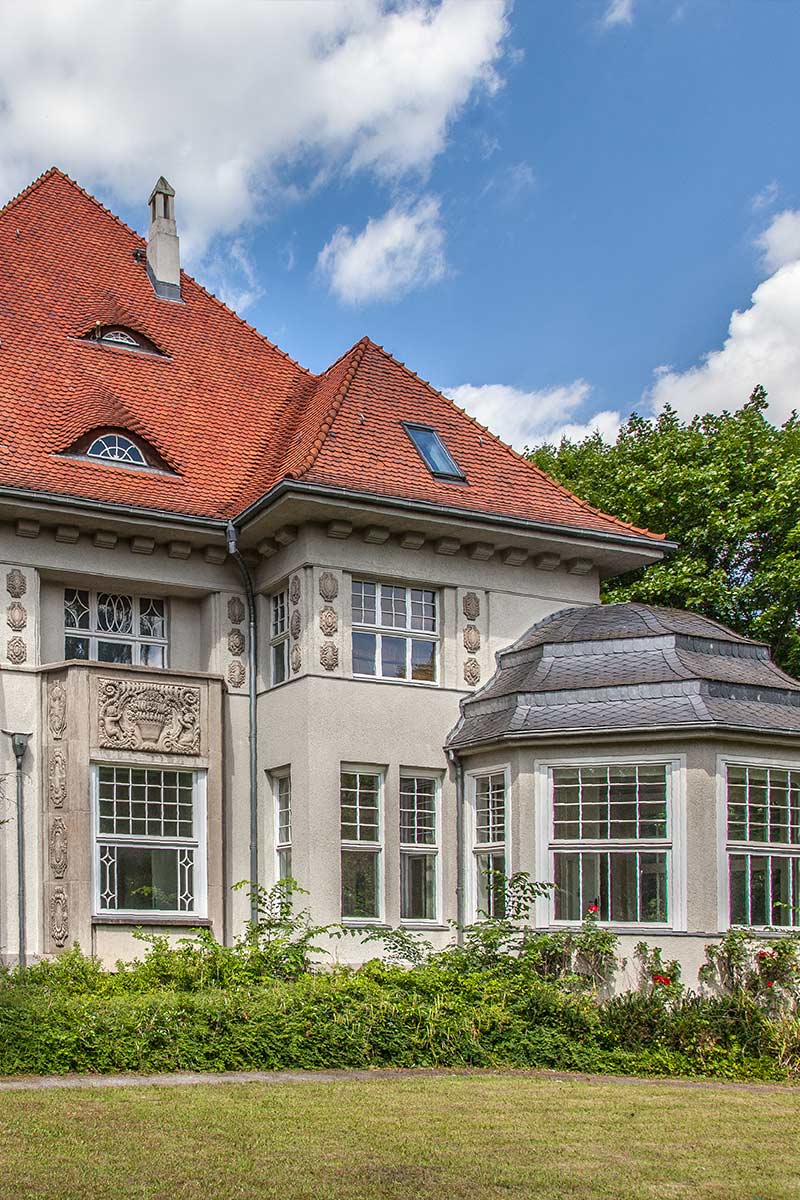Historische Villa mit roten Ziegeldach, großen Fenstern und dekorativen Fassadenelementen.
