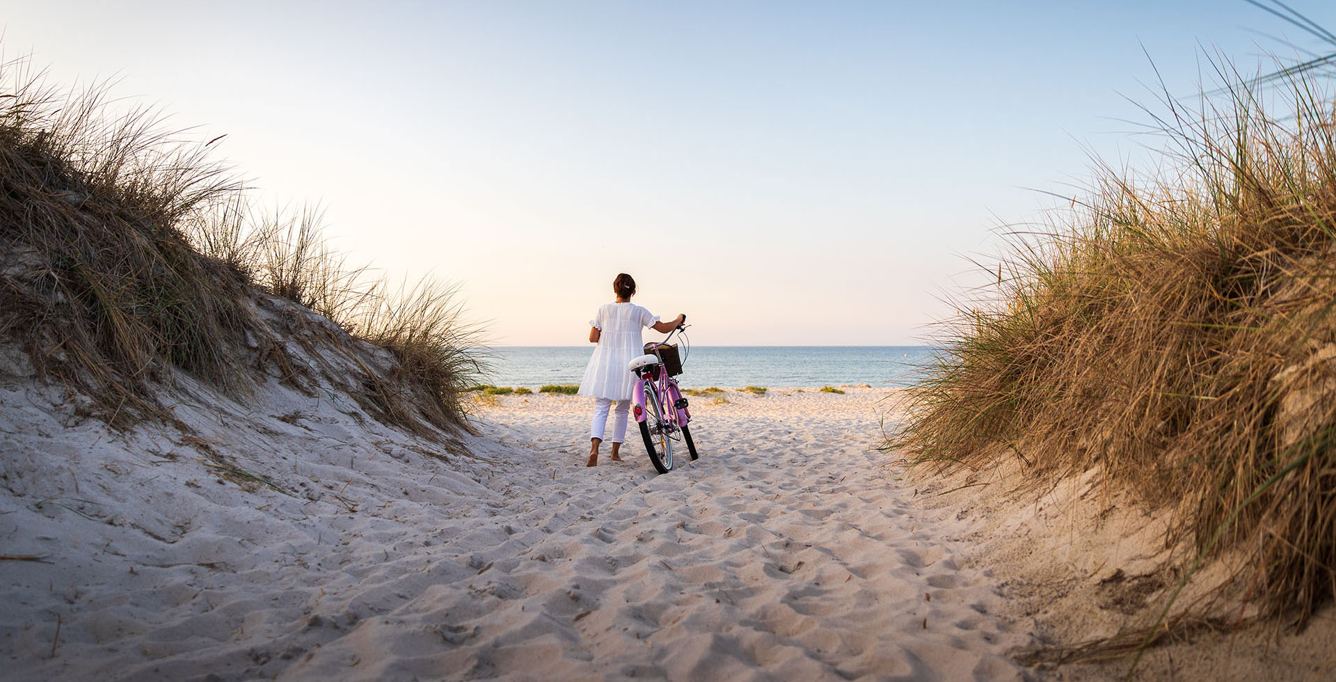 Frau in weißem Kleid mit Fahrrad auf sandigem Pfad zwischen Dünen mit Blick auf die ruhige Ostsee im Urlaub auf Rügen.