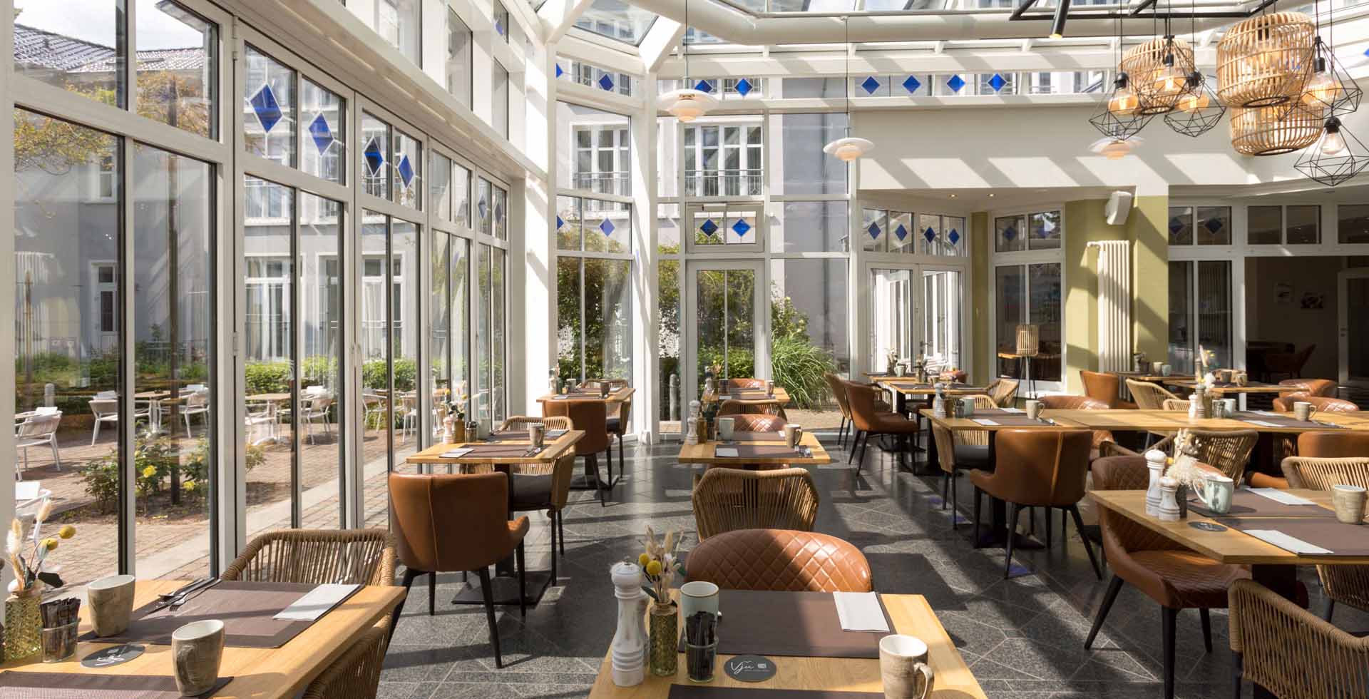 Gemütlicher Wintergarten des Vju Hotels Rügen mit Frühstückstischen und großen Fenstern.