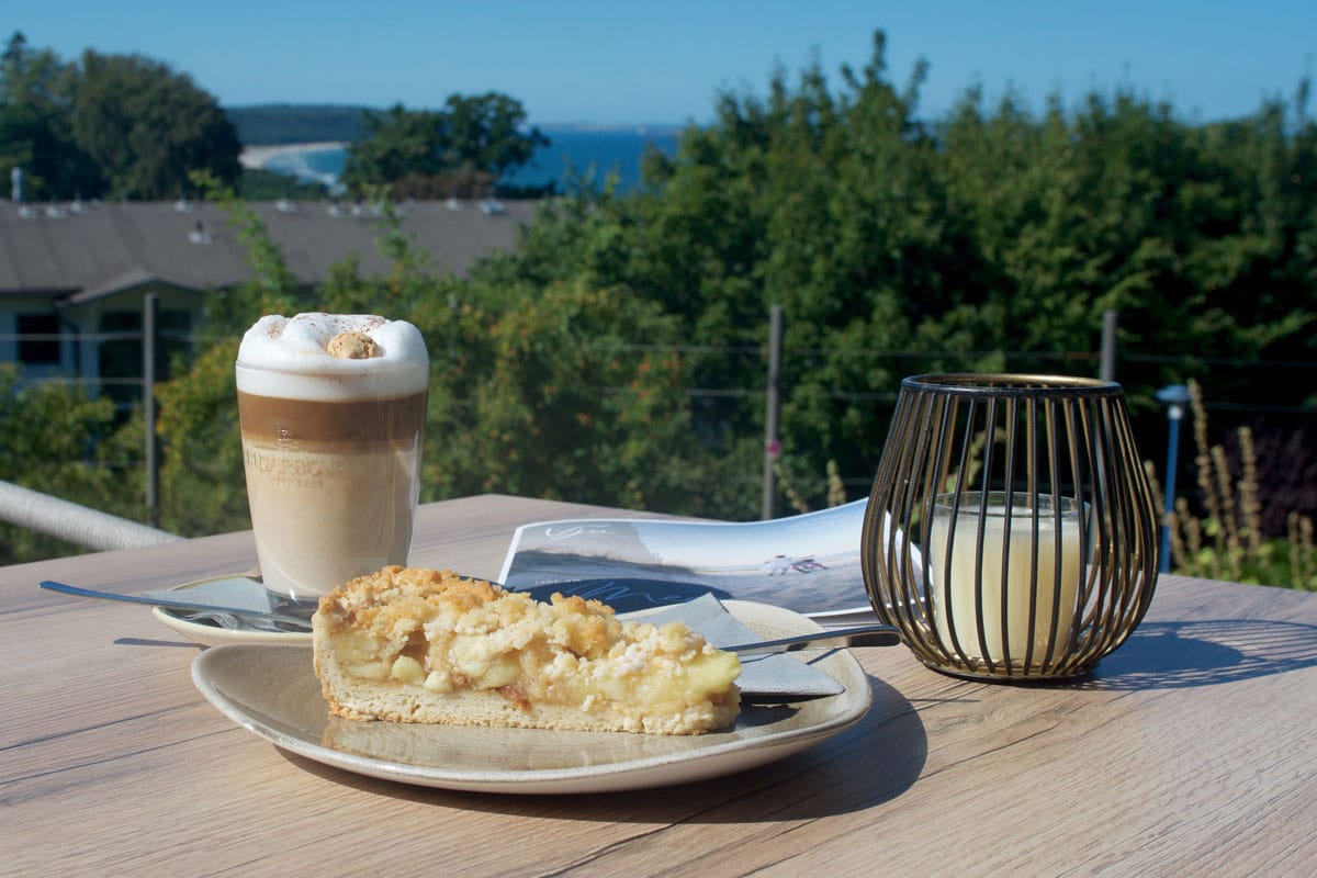 Ein Latte Macchiato und ein Stück Apfelkuchen auf einem Tisch mit Ausblick auf die Ostsee auf der See-Terasse im Vju Hotel.