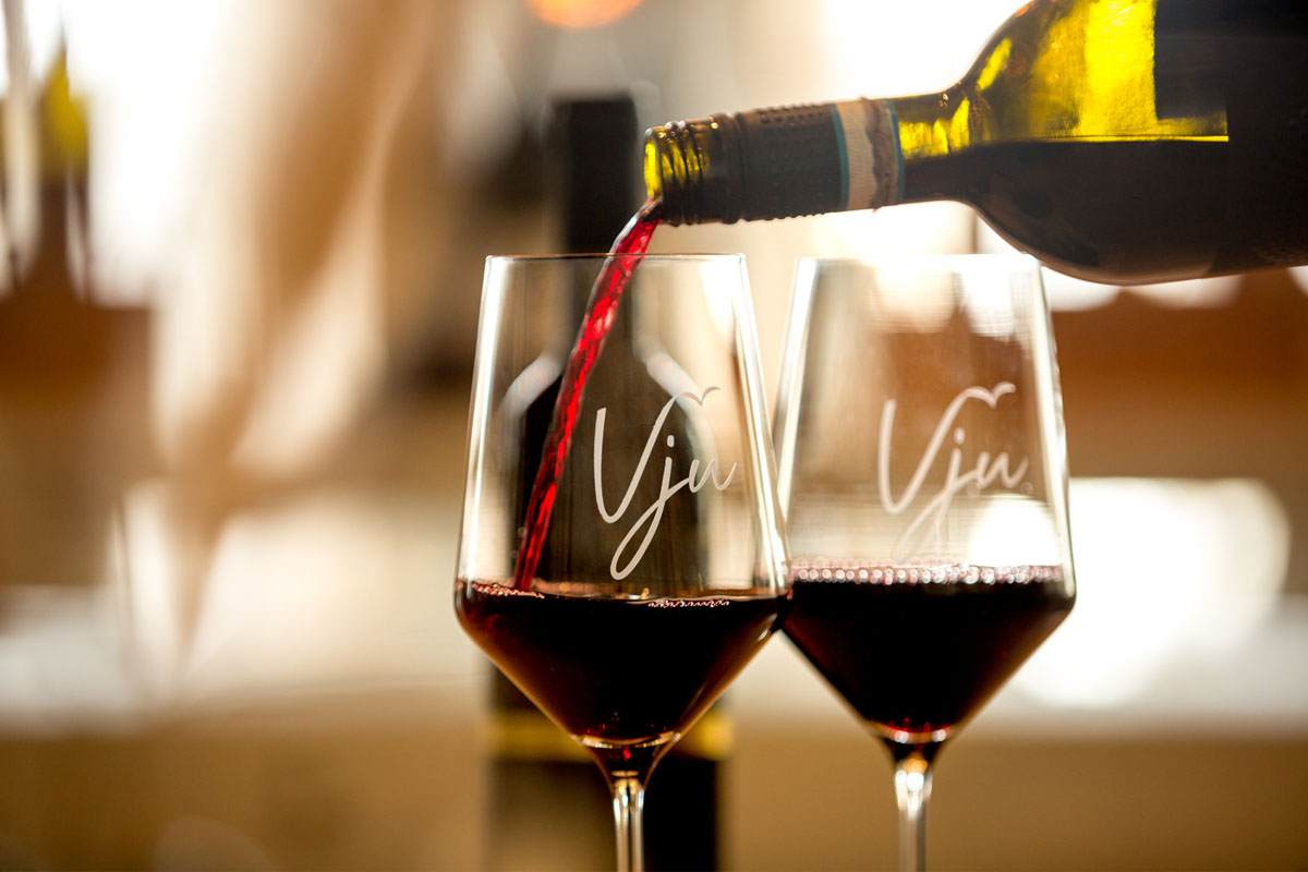 Nahaufnahme von zwei Gläsern Rotwein, eines wird gerade eingeschenkt, mit dem Schriftzug Vju im Strandläufer auf Rügen.