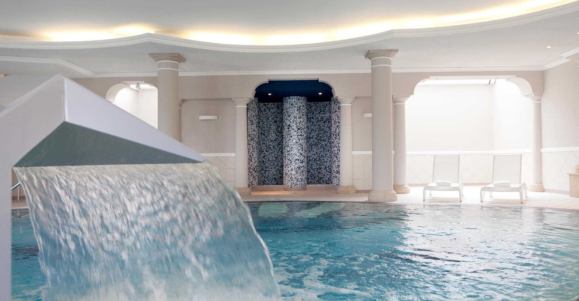 Großzügiger Indoorpool im Vju Hotel auf Rügen, ausgestattet mit Wasserfall, Liegen und stilvoller Inneneinrichtung für Erholung.