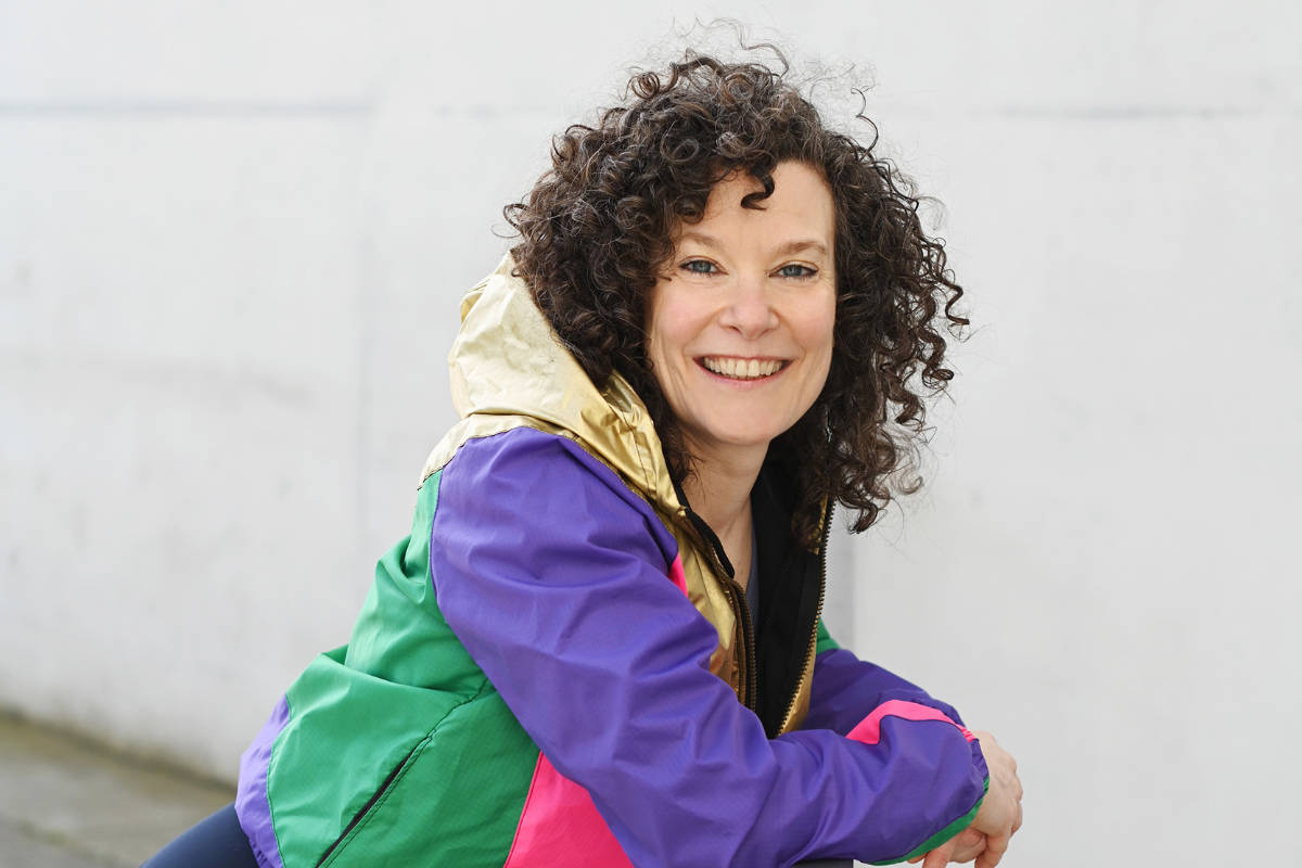 Yogalehrerin Barbara Pascaly sitzt lächelnd vor einer hellen Wand und trägt eine farbenfrohe Jacke bereit für Yoga auf Rügen.