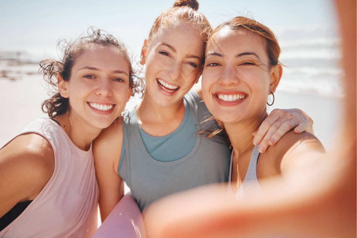 Drei junge Frauen am Strand in Yogakleidung machen ein Selfie.