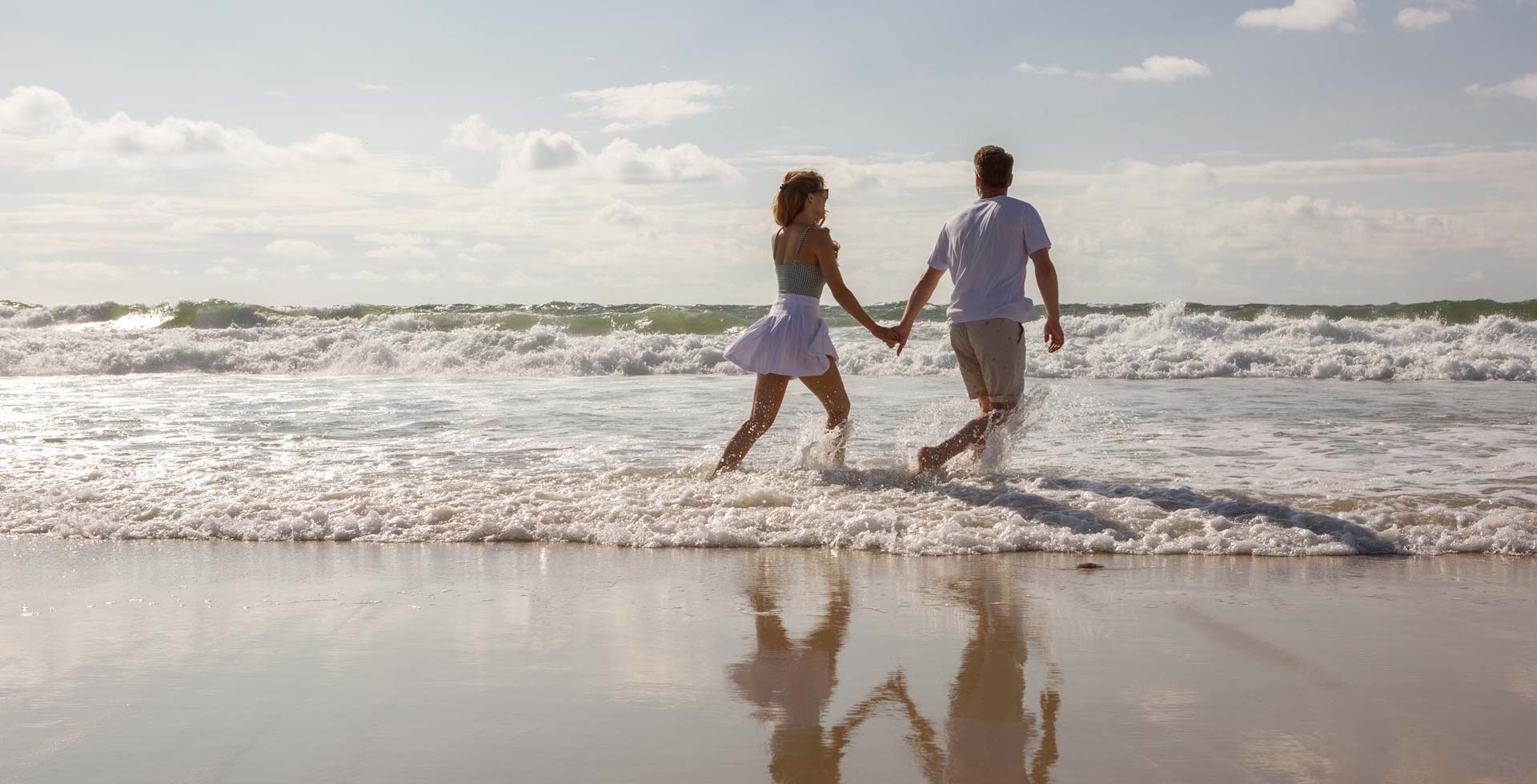 Ein Paar läuft Hand in Hand am Sandstrand vor dem Wyn. Strandhotel Sylt entlang, umgeben von Wellen und Sonne.