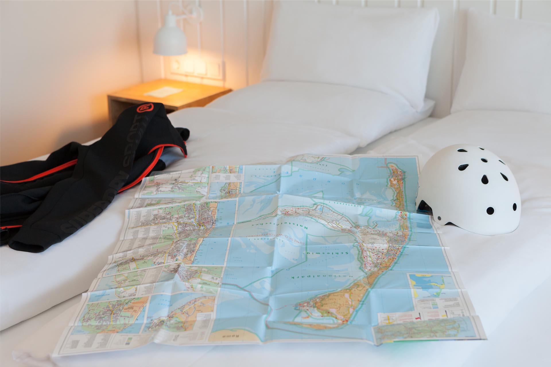 Eine Karte von Sylt liegt auf einem Bett im Wyn Strandhotel Sylt, daneben ein Fahrradhelm und eine Jacke.